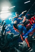 A figure of a superhero in a dynamic pose, featuring a clenched fist and an intense expression, stands against a backdrop with bold colors and graphics. The figure's costume is blue with a red cape, and there's a sense of motion and power in the scene. Dark, rugged terrain appears in the foreground, adding depth.