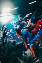 A figure of a superhero in a dynamic pose, featuring a clenched fist and an intense expression, stands against a backdrop with bold colors and graphics. The figure's costume is blue with a red cape, and there's a sense of motion and power in the scene. Dark, rugged terrain appears in the foreground, adding depth.