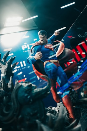 A figure of a superhero in a dynamic pose, featuring a clenched fist and an intense expression, stands against a backdrop with bold colors and graphics. The figure's costume is blue with a red cape, and there's a sense of motion and power in the scene. Dark, rugged terrain appears in the foreground, adding depth.