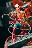 A detailed action figure of a character wearing a red and green costume, featuring a helmet with protruding horns and a chain weapon that wraps around their body. The figure is posed dynamically, standing on a rocky base. The background includes a mix of colors and possibly other figures or decorations, adding to the dramatic effect.