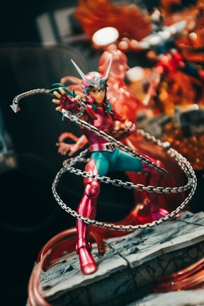 A detailed action figure of a character wearing a red and green costume, featuring a helmet with protruding horns and a chain weapon that wraps around their body. The figure is posed dynamically, standing on a rocky base. The background includes a mix of colors and possibly other figures or decorations, adding to the dramatic effect.