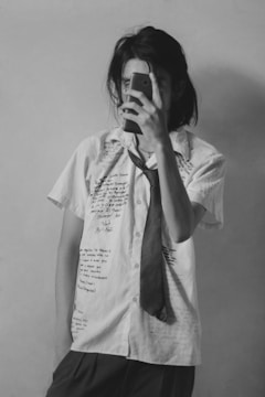 A person is standing against a plain background, taking a selfie with a smartphone. They are wearing a short-sleeved white shirt covered with handwritten text in various parts and a loosely tied necktie. The overall look is casual and somewhat artistic.