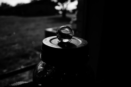 A black and white photograph highlighting a glass marble placed on top of a jar lid. The background is blurred, showcasing an outdoor environment with faint outlines of trees and grass. The play of light and shadows creates a contrast between the shiny, reflective surface of the marble and the dark surroundings.