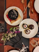 A table setting with a rich chocolate cake adorned with a red ribbon and a gold base on one plate, alongside two smaller desserts with chocolate garnish and cherry toppings on another plate. There is a bouquet of vibrant flowers, including purple, white, and green blooms, wrapped in black paper. Several metallic spoons are arranged nearby, with a glass partially filled with a liquid.