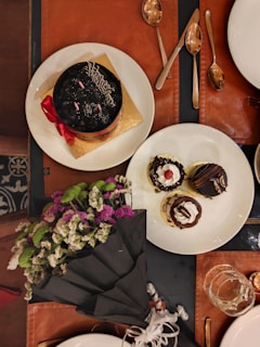 A table setting with a rich chocolate cake adorned with a red ribbon and a gold base on one plate, alongside two smaller desserts with chocolate garnish and cherry toppings on another plate. There is a bouquet of vibrant flowers, including purple, white, and green blooms, wrapped in black paper. Several metallic spoons are arranged nearby, with a glass partially filled with a liquid.