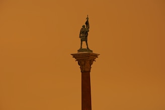 A statue of a warrior holding a flag stands atop a tall column against a monochromatic orange sky.