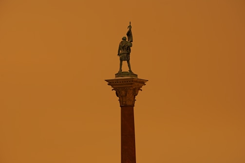 A statue of a warrior holding a flag stands atop a tall column against a monochromatic orange sky.