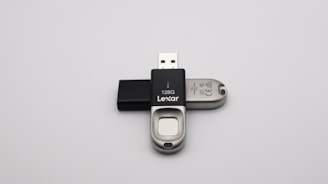 A 128GB Lexar USB flash drive with a metal and black plastic design is positioned against a plain grey background. The flash drive has a distinctive sliding mechanism and branding visible on the black part.