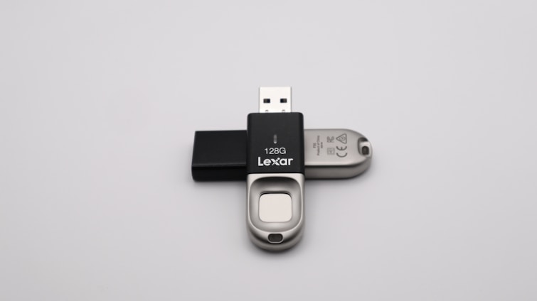 A 128GB Lexar USB flash drive with a metal and black plastic design is positioned against a plain grey background. The flash drive has a distinctive sliding mechanism and branding visible on the black part.
