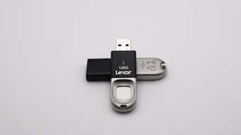 A 128GB Lexar USB flash drive with a metal and black plastic design is positioned against a plain grey background. The flash drive has a distinctive sliding mechanism and branding visible on the black part.