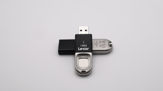 A 128GB Lexar USB flash drive with a metal and black plastic design is positioned against a plain grey background. The flash drive has a distinctive sliding mechanism and branding visible on the black part.