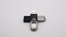 A 128GB Lexar USB flash drive with a metal and black plastic design is positioned against a plain grey background. The flash drive has a distinctive sliding mechanism and branding visible on the black part.