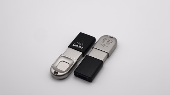 Two USB flash drives are positioned on a plain, light-colored surface. One drive is a Lexar model with 128GB capacity, featuring a slide mechanism with a metallic and black exterior. The other drive has a black cap and metallic body, with identifiable symbols and text inscribed.