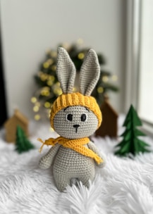 A crocheted grey bunny wearing a yellow hat and scarf stands on a plush white surface. In the background, there are small Christmas trees and decorative wooden houses with out-of-focus festive lights.