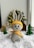 A crocheted grey bunny wearing a yellow hat and scarf stands on a plush white surface. In the background, there are small Christmas trees and decorative wooden houses with out-of-focus festive lights.