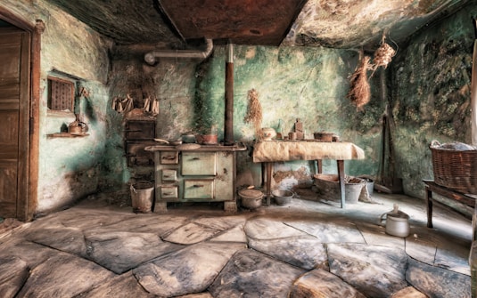 An old rustic kitchen with weathered green walls and a stone floor. A vintage stove with pots and pans is against the wall, surrounded by various kitchen utensils and dried herbs hanging from the ceiling. A wooden table with a cloth draped over it holds more cooking items. The room is dimly lit, evoking a sense of nostalgia.