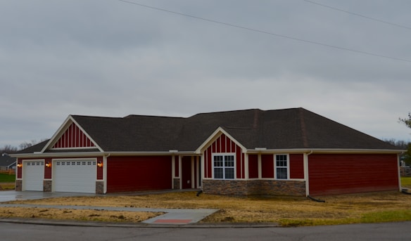 A single-story house painted in red with white trim, featuring a large three-car garage and multiple windows. The roof is dark grey, and there is a stone veneer accent at the base of the walls. The house is situated on a grassy lot with a driveway and sidewalk out front under an overcast sky.