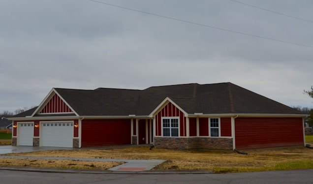 A single-story house painted in red with white trim, featuring a large three-car garage and multiple windows. The roof is dark grey, and there is a stone veneer accent at the base of the walls. The house is situated on a grassy lot with a driveway and sidewalk out front under an overcast sky.
