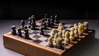 A chessboard with intricately carved pieces is set up for a game. The chess pieces are uniquely styled, with one side featuring onyx black pieces and the other side showcasing ivory white pieces. The board itself appears to be made of a combination of dark and light marble squares bordered by wood. It is placed on a smooth, dark surface with a dimly lit background.