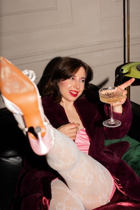 A person is lounging on a couch wearing a burgundy robe and patterned white stockings. They are holding a glass of champagne and appear to be in a celebratory mood, smiling with bright red lipstick. Their foot is playfully extended towards the camera, showcasing a high-heeled shoe.
