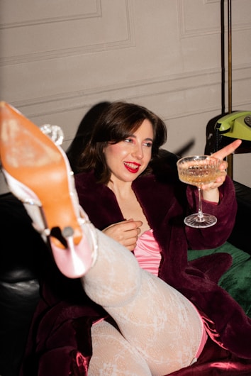 A person is lounging on a couch wearing a burgundy robe and patterned white stockings. They are holding a glass of champagne and appear to be in a celebratory mood, smiling with bright red lipstick. Their foot is playfully extended towards the camera, showcasing a high-heeled shoe.