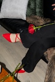 A pair of crossed legs wearing black pants and bright red shoes is visible. A red rose lies across the legs, with its petals and stem clearly visible. The background includes a green velvet pillow, a white pillow, and a dark couch.