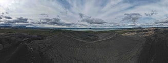 A vast volcanic landscape with a prominent crater in the center. The surrounding terrain is mostly dark and rocky, with patches of green vegetation in the distance. The horizon features a range of mountains under a cloudy sky, creating a dramatic and expansive view.