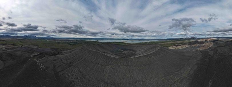 A vast volcanic landscape with a prominent crater in the center. The surrounding terrain is mostly dark and rocky, with patches of green vegetation in the distance. The horizon features a range of mountains under a cloudy sky, creating a dramatic and expansive view.