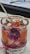 A glass filled with a red gel-like substance features small seashells embedded within. There is a purple flower petal partially submerged in the gel. A piece of twine is tied around the glass, and a wooden stick rests on top, secured with a knotted string.