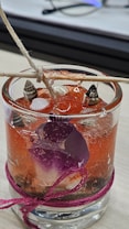A glass filled with a red gel-like substance features small seashells embedded within. There is a purple flower petal partially submerged in the gel. A piece of twine is tied around the glass, and a wooden stick rests on top, secured with a knotted string.