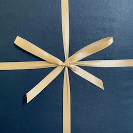 A neatly tied golden ribbon is placed against a dark blue, textured background. The ribbon has a glossy finish that catches the light, and is tied in a symmetrical bow with long, smooth tails crossing over each other.