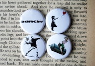 Four round badges are placed on top of a page with text. Each badge features iconic artworks. The top left badge has the word 'BANKSY' in bold, stylized lettering. The top right badge depicts a silhouette of a girl releasing a red heart-shaped balloon. The bottom left one shows a figure in a spray-painted style, appearing to throw a bouquet of flowers. The bottom right badge portrays a retro-style image of a woman sitting on a large canister, reminiscent of classic pin-up art.