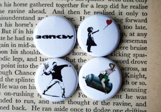 Four round badges are placed on top of a page with text. Each badge features iconic artworks. The top left badge has the word 'BANKSY' in bold, stylized lettering. The top right badge depicts a silhouette of a girl releasing a red heart-shaped balloon. The bottom left one shows a figure in a spray-painted style, appearing to throw a bouquet of flowers. The bottom right badge portrays a retro-style image of a woman sitting on a large canister, reminiscent of classic pin-up art.