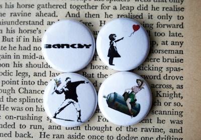 Four round badges are placed on top of a page with text. Each badge features iconic artworks. The top left badge has the word 'BANKSY' in bold, stylized lettering. The top right badge depicts a silhouette of a girl releasing a red heart-shaped balloon. The bottom left one shows a figure in a spray-painted style, appearing to throw a bouquet of flowers. The bottom right badge portrays a retro-style image of a woman sitting on a large canister, reminiscent of classic pin-up art.