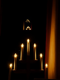 A series of electric candle lights arranged in a candelabra pattern emits a warm glow. The light reflects in a dark, mirrored surface, creating multiple iterations of the candlelights stretching into the background. Surrounding the candles is a dark room, giving prominence to the bright lights.