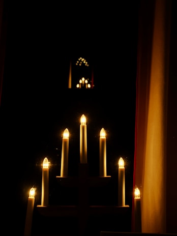 A series of electric candle lights arranged in a candelabra pattern emits a warm glow. The light reflects in a dark, mirrored surface, creating multiple iterations of the candlelights stretching into the background. Surrounding the candles is a dark room, giving prominence to the bright lights.