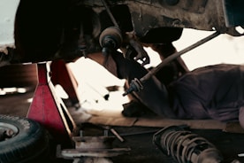 A mechanic is lying on the ground under a vehicle, working on its suspension system. The vehicle is supported by a red hydraulic jack stand. Various car parts and a tire are scattered nearby on the concrete floor, suggesting a repair or maintenance activity in a workshop environment.
