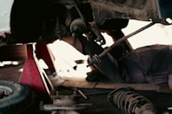 A mechanic is lying on the ground under a vehicle, working on its suspension system. The vehicle is supported by a red hydraulic jack stand. Various car parts and a tire are scattered nearby on the concrete floor, suggesting a repair or maintenance activity in a workshop environment.