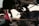 A mechanic is lying on the ground under a vehicle, working on its suspension system. The vehicle is supported by a red hydraulic jack stand. Various car parts and a tire are scattered nearby on the concrete floor, suggesting a repair or maintenance activity in a workshop environment.