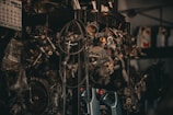 A collection of various metal car parts and mechanical components hangs in a cluttered garage-like setting. The scene includes coiled hoses, engine parts, and oil containers. The lighting is dim, creating a gritty, industrial atmosphere.