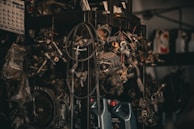 A collection of various metal car parts and mechanical components hangs in a cluttered garage-like setting. The scene includes coiled hoses, engine parts, and oil containers. The lighting is dim, creating a gritty, industrial atmosphere.