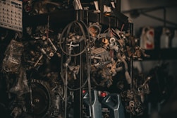 A collection of various metal car parts and mechanical components hangs in a cluttered garage-like setting. The scene includes coiled hoses, engine parts, and oil containers. The lighting is dim, creating a gritty, industrial atmosphere.