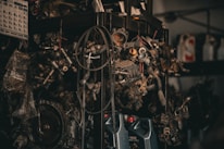 A collection of various metal car parts and mechanical components hangs in a cluttered garage-like setting. The scene includes coiled hoses, engine parts, and oil containers. The lighting is dim, creating a gritty, industrial atmosphere.