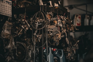 A collection of various metal car parts and mechanical components hangs in a cluttered garage-like setting. The scene includes coiled hoses, engine parts, and oil containers. The lighting is dim, creating a gritty, industrial atmosphere.