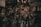 A collection of various metal car parts and mechanical components hangs in a cluttered garage-like setting. The scene includes coiled hoses, engine parts, and oil containers. The lighting is dim, creating a gritty, industrial atmosphere.