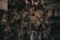A collection of various metal car parts and mechanical components hangs in a cluttered garage-like setting. The scene includes coiled hoses, engine parts, and oil containers. The lighting is dim, creating a gritty, industrial atmosphere.