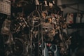 A collection of various metal car parts and mechanical components hangs in a cluttered garage-like setting. The scene includes coiled hoses, engine parts, and oil containers. The lighting is dim, creating a gritty, industrial atmosphere.