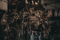 A collection of various metal car parts and mechanical components hangs in a cluttered garage-like setting. The scene includes coiled hoses, engine parts, and oil containers. The lighting is dim, creating a gritty, industrial atmosphere.
