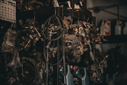 A collection of various metal car parts and mechanical components hangs in a cluttered garage-like setting. The scene includes coiled hoses, engine parts, and oil containers. The lighting is dim, creating a gritty, industrial atmosphere.