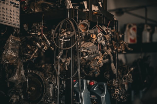 A collection of various metal car parts and mechanical components hangs in a cluttered garage-like setting. The scene includes coiled hoses, engine parts, and oil containers. The lighting is dim, creating a gritty, industrial atmosphere.